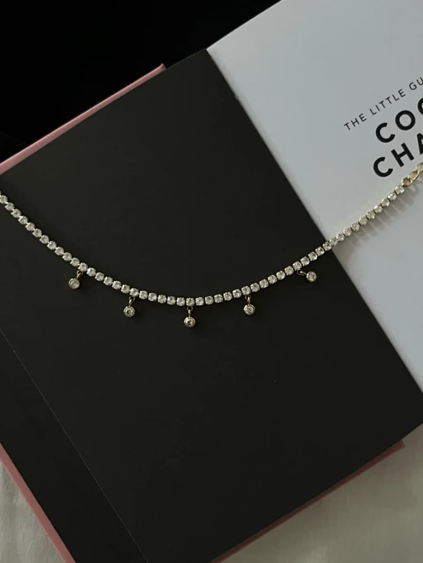 A gold-tone bracelet with a continuous chain of clear crystal-like stones and several dangling smaller clear charms, draped over an open book.
