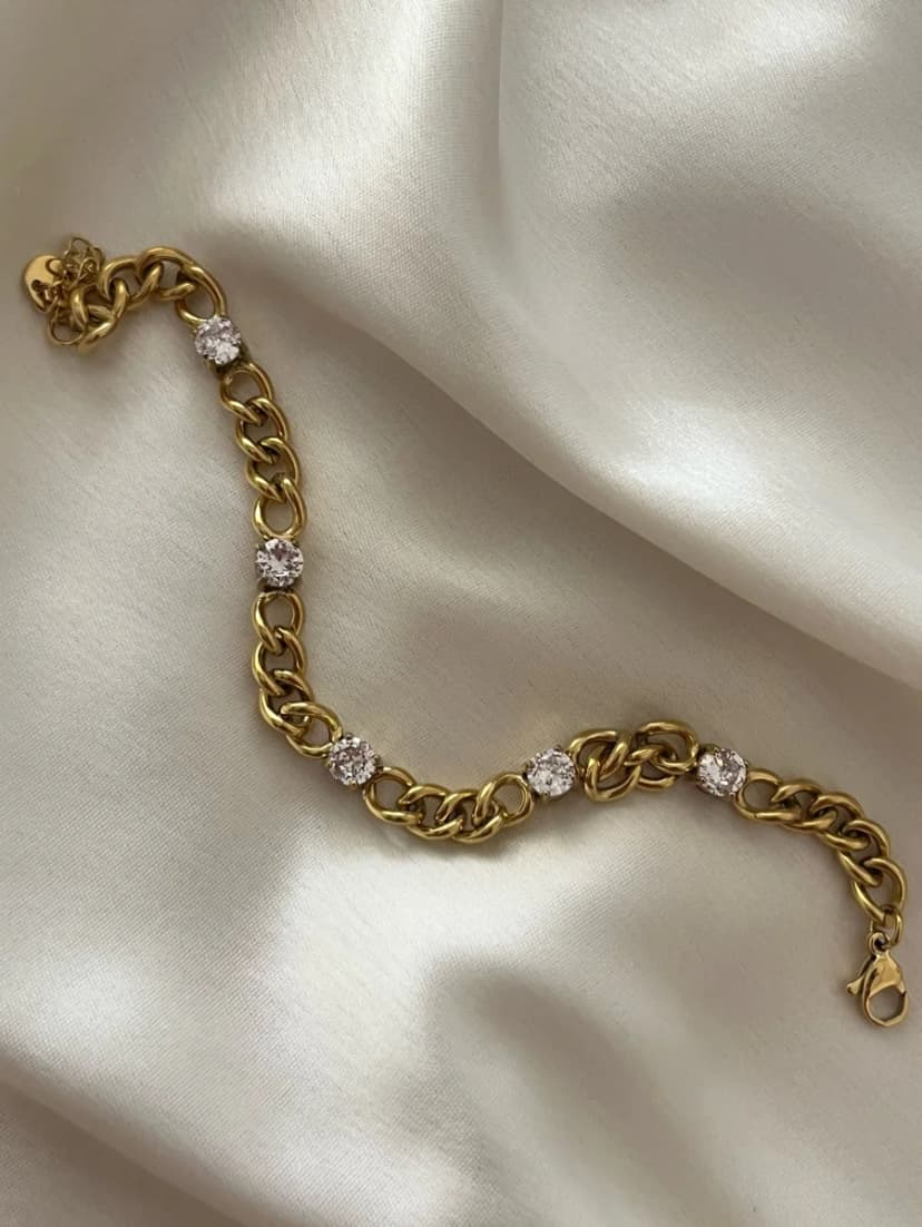 A chunky gold-tone curb chain bracelet with five round crystal stations, laid on white satin fabric.