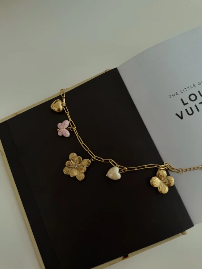 A gold-tone chunky chain bracelet with a puffy heart, pink butterfly, two textured flower, and a pearl-like bead charms, on an open book.
