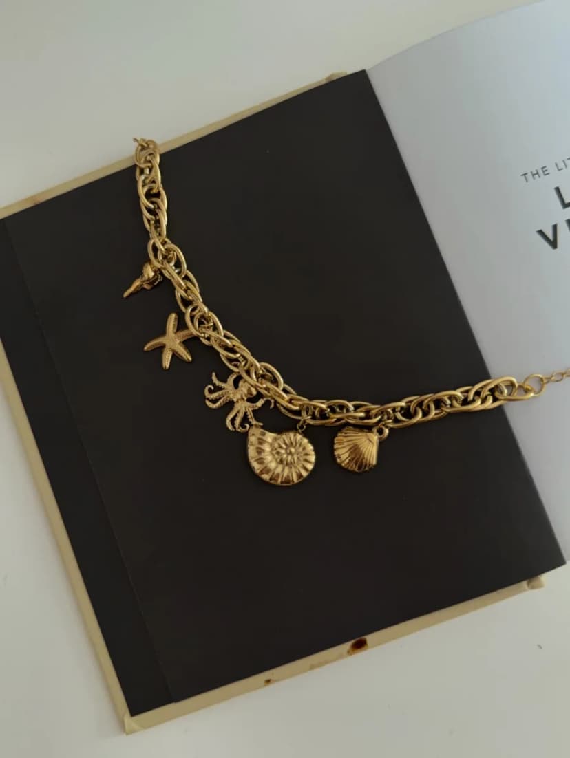 A gold-tone chunky chain bracelet with a cone shell, starfish, octopus, spiral shell, and scallop shell charms, on an open book.
