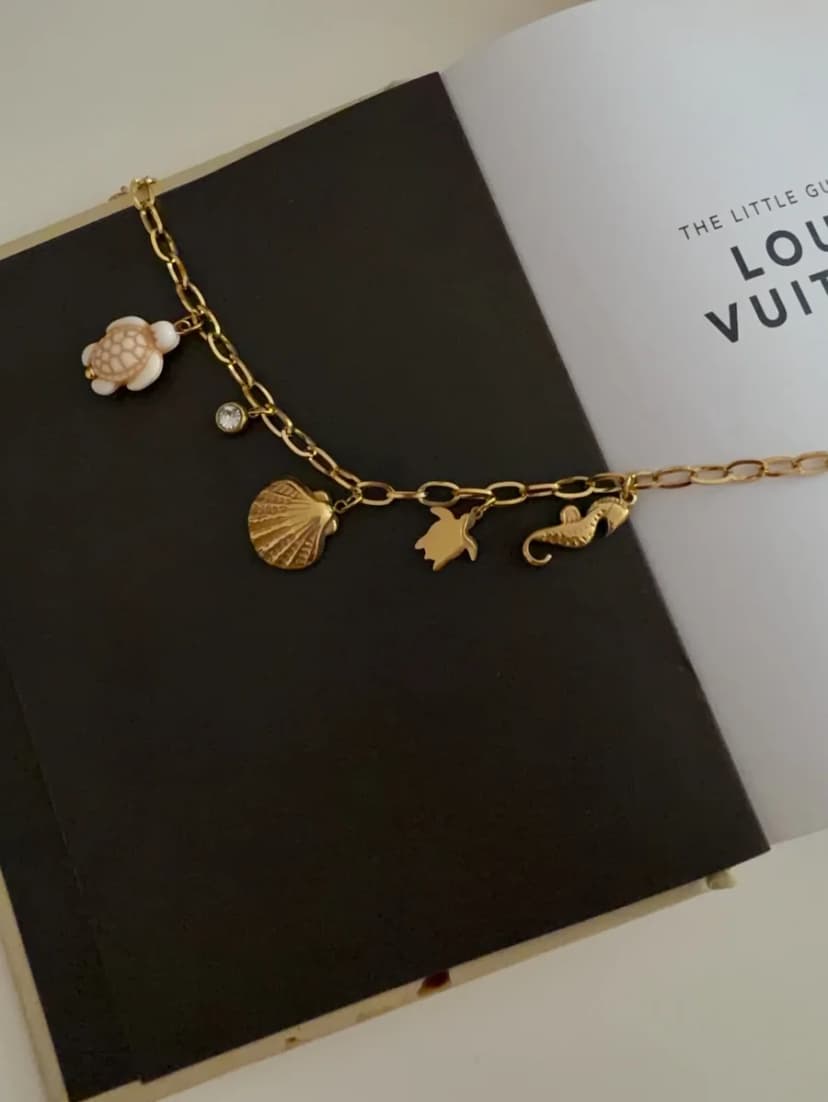 A gold-tone chunky chain bracelet with a turtle, clear crystal, seashell, starfish, and seahorse charms, on an open book.